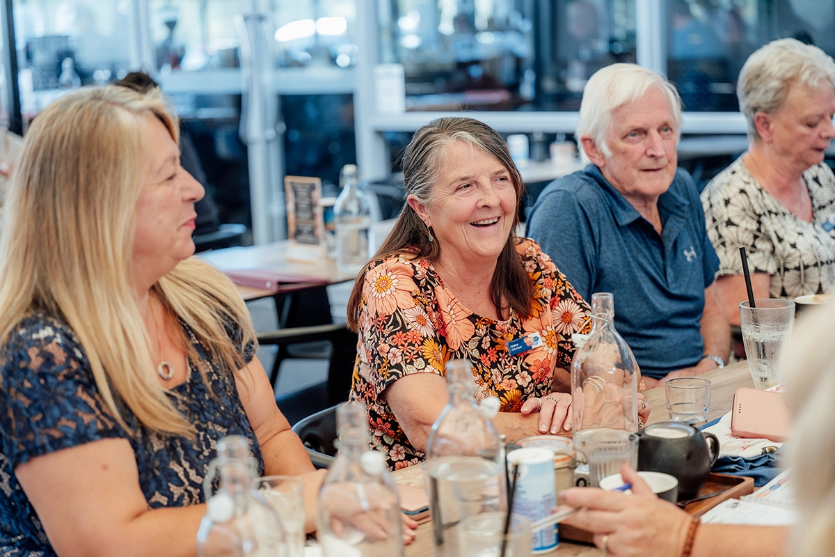 A group of older adults sit together at a Talking Café, smiling and talking. The table is covered with glasses and cups, and the warm, friendly atmosphere encourages everyone to meet new people whilst Living Our Best Life.